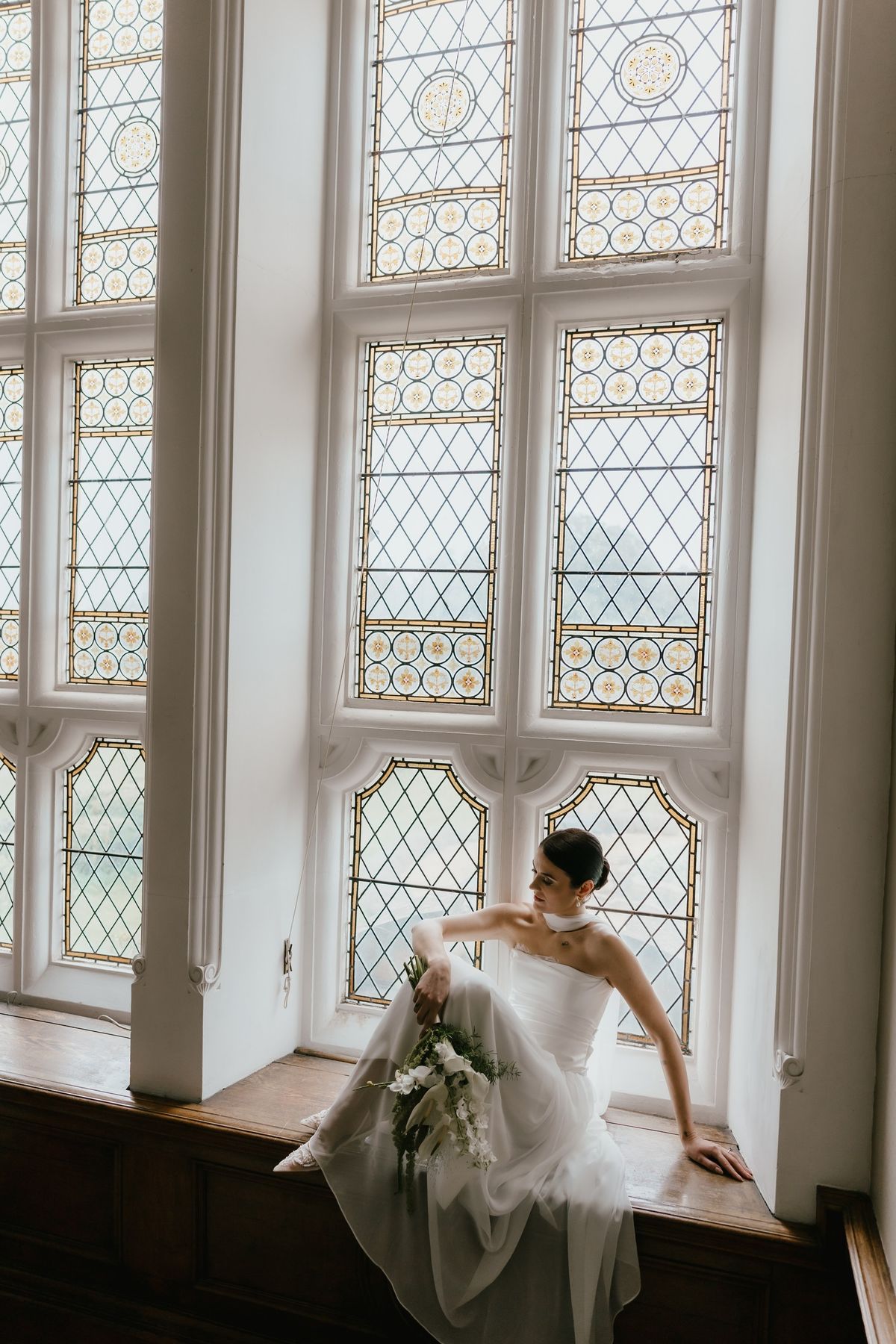 Bride in a lace edged satin corset and gathered silk chiffon skirt seated beneath leaded windows