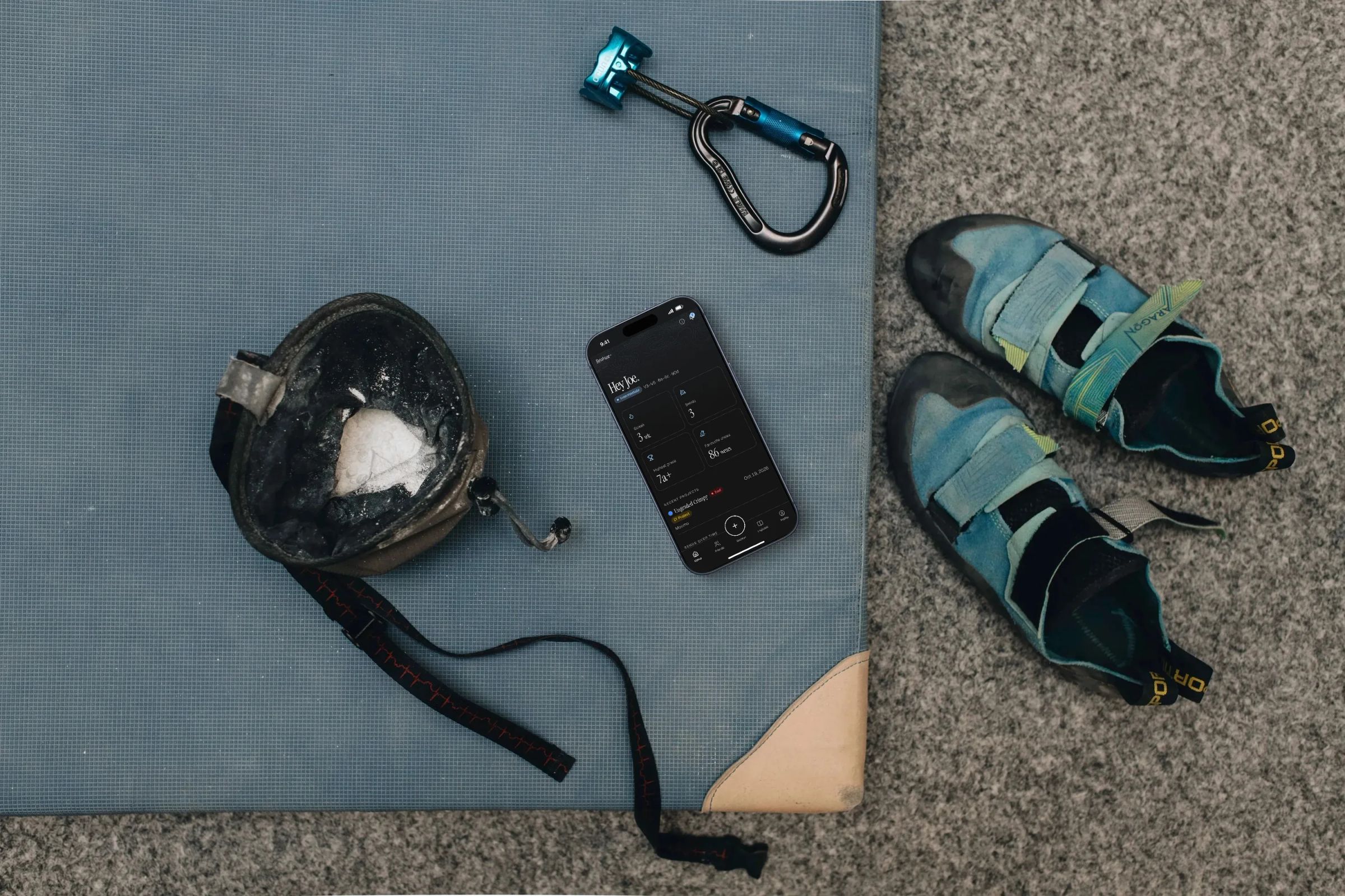 The Betabase+ home screen on a phone lying among climbing gear: shoes, a chalk bag and a carabiner