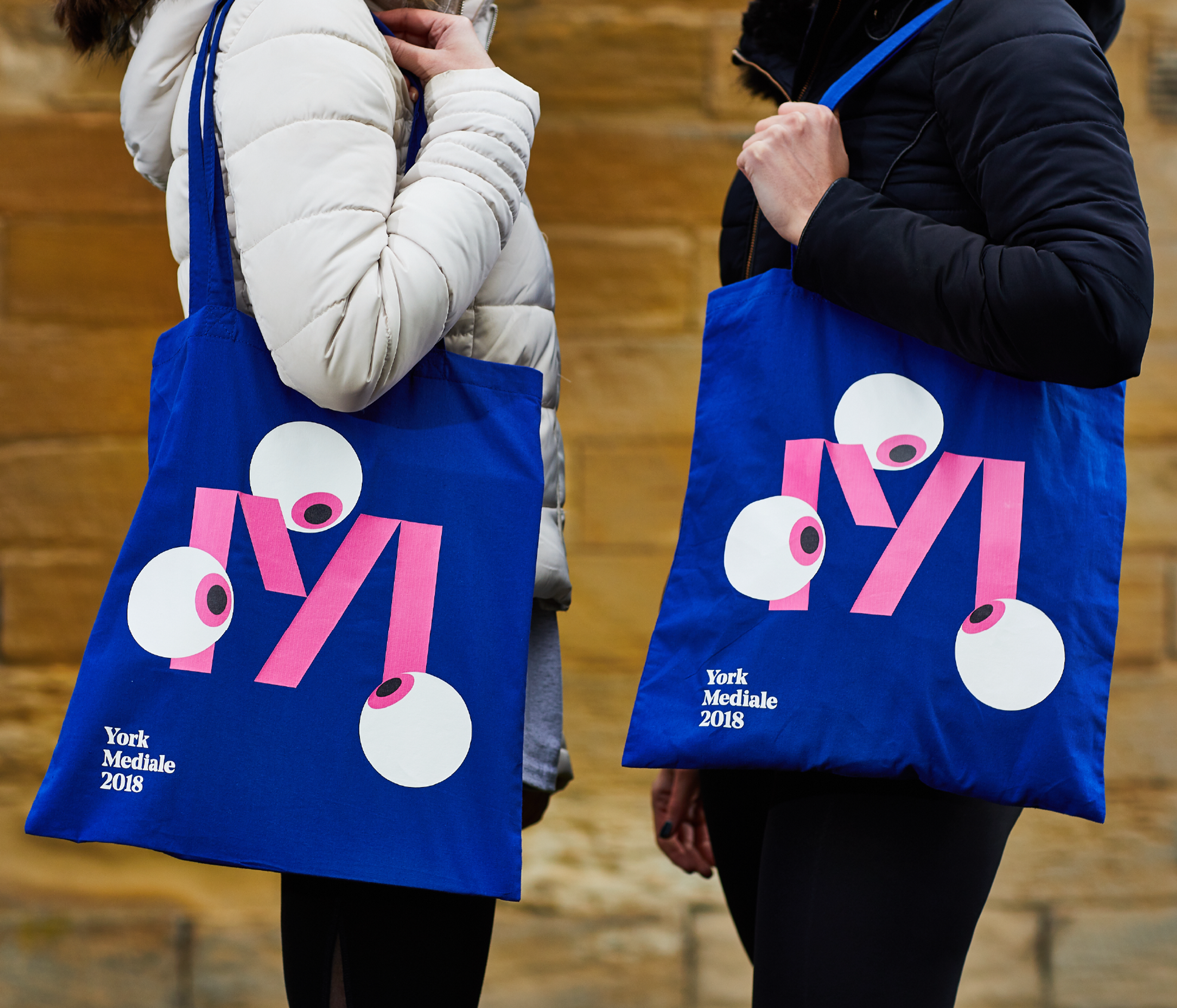 Two people wearing winter puffer coats hold electric blue tote bags with the Mediale 'M' logo. Three eye balls surround the M, as if looking at it.