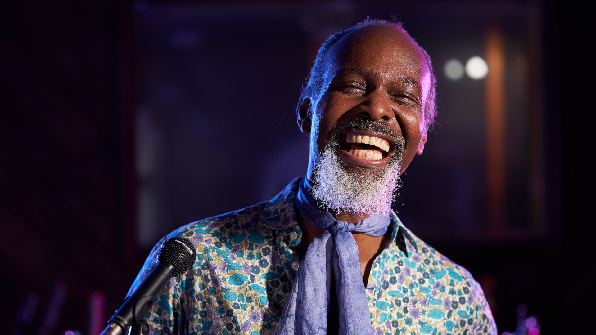Elderly man wearing a funky shirt and knotted scarf is photographed mid laugh. The background setting is dark with purple lighting, that makes it look like it was taken from inside a club of some sorts. We are purely focussed on the man, as he's shot with a deep depth of focus