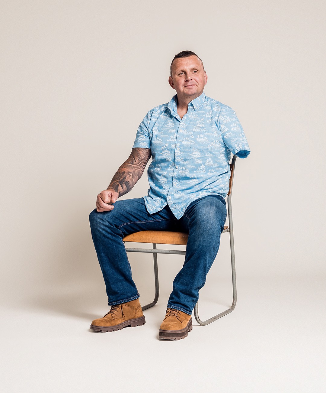 An amputee sits on a simple chair, with a cream coloured background. He wears a walm smile, and blue hawaiian shirt
