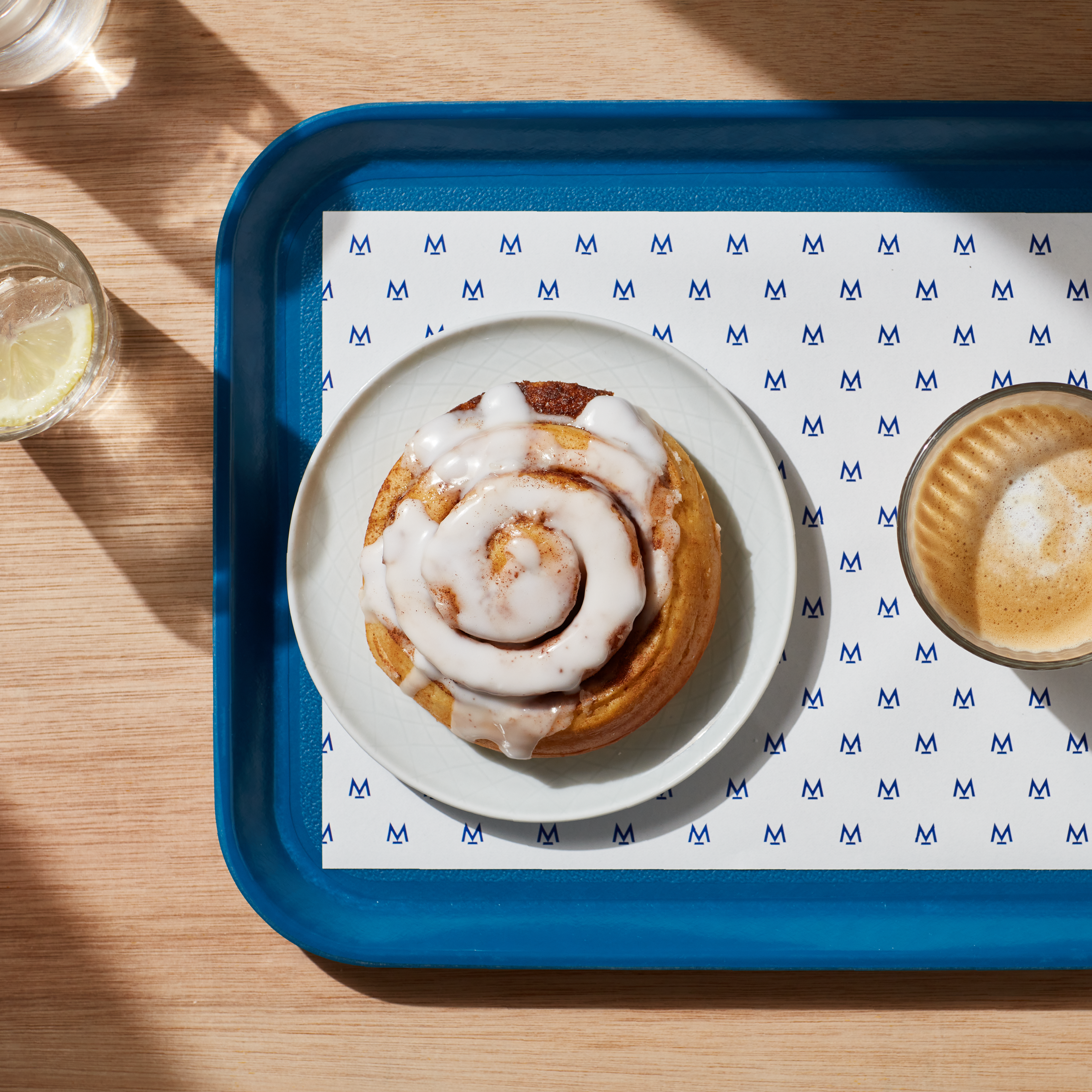 Woodern scandi style table, with a cup of water and lemon on the left. There is a blue tray on the right. On the tray is a sheet of Modern Standard Branding paper, cinnamon roll and accompanying coffee in a glass cup