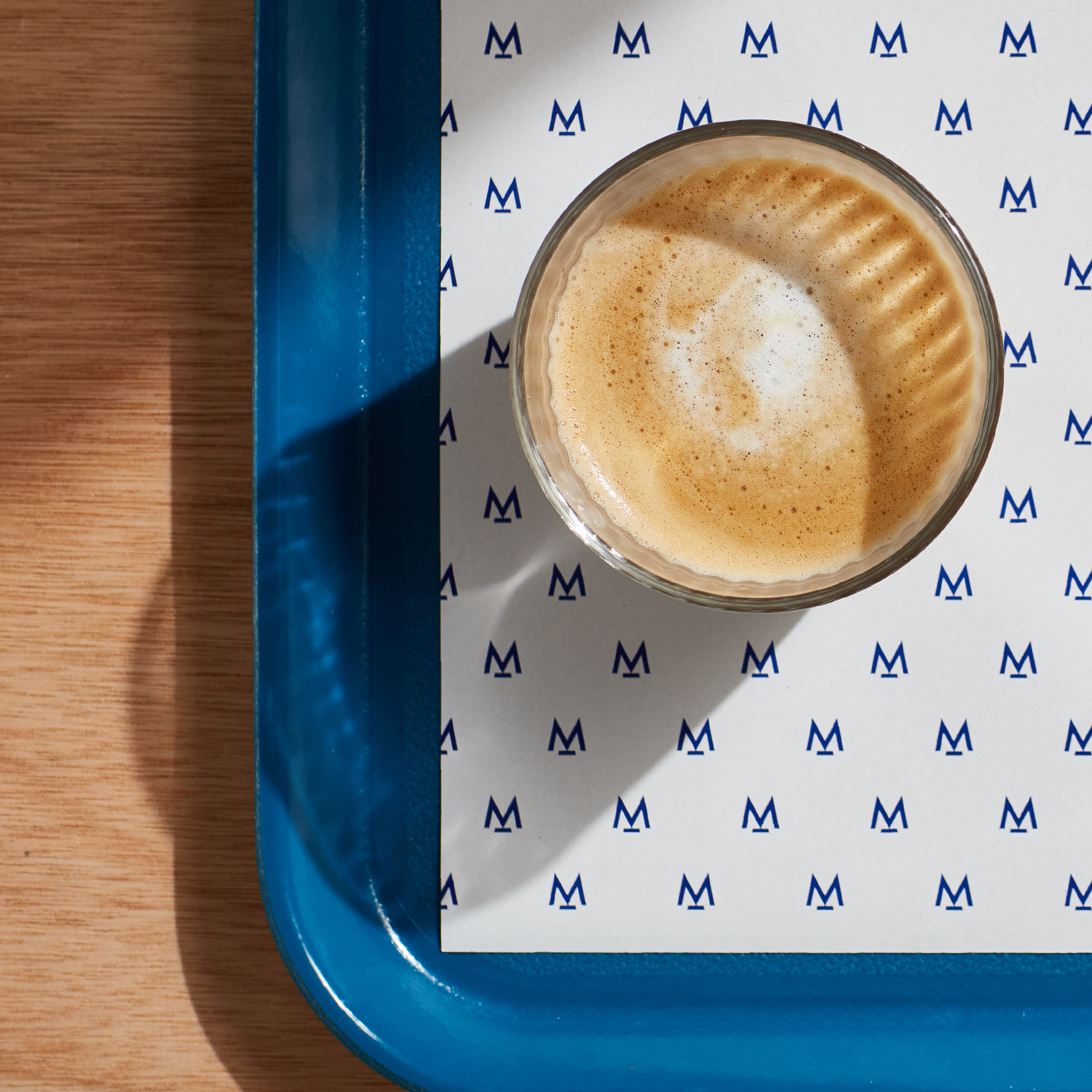 Woodern scandi style table is seen on the left, and a blue tray with a branding sheet of paper placed on the right. On this tray we have a freshly brewed coffee in a glass cup