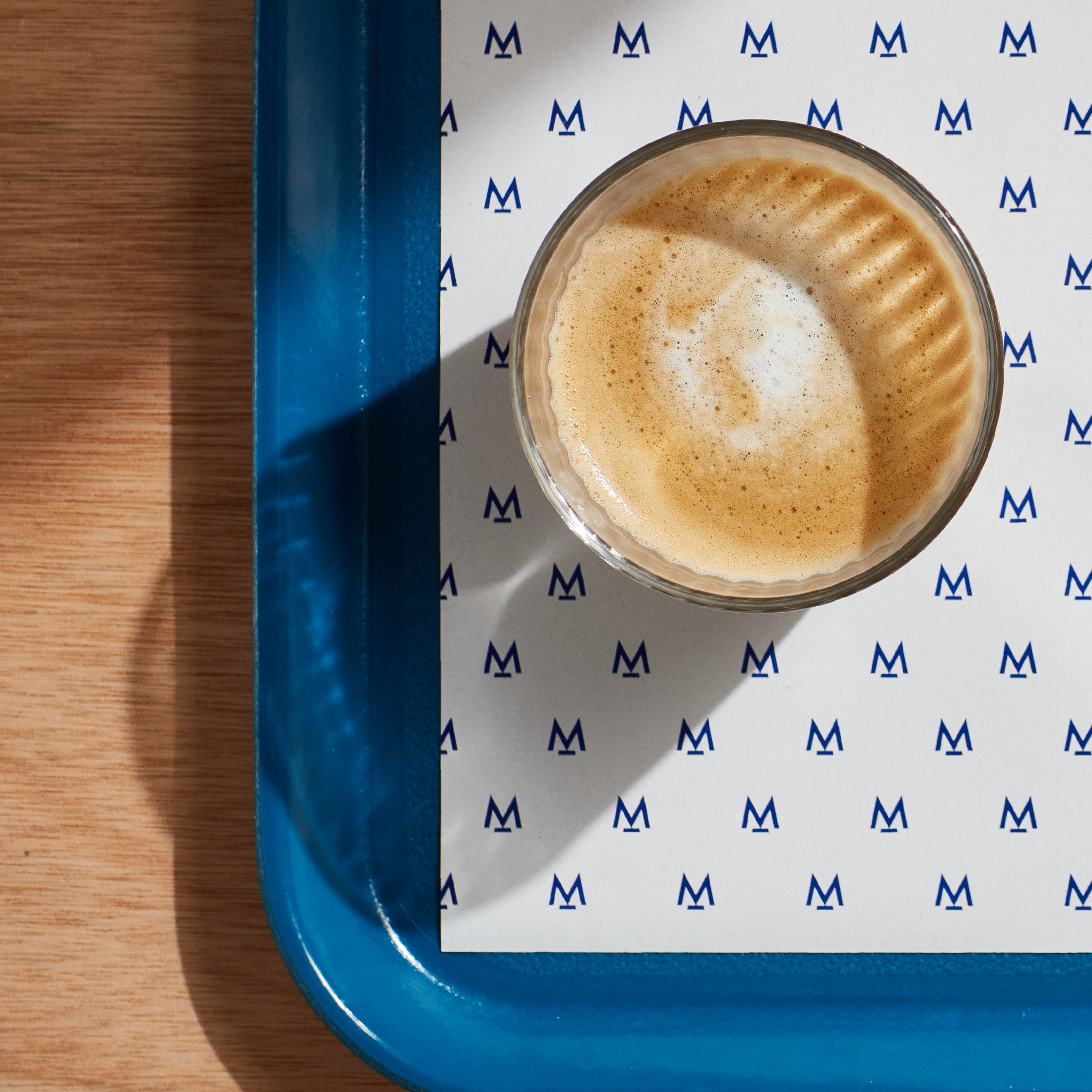 Woodern scandi style table is seen on the left, and a blue tray with a branding sheet of paper placed on the right. On this tray we have a freshly brewed coffee in a glass cup