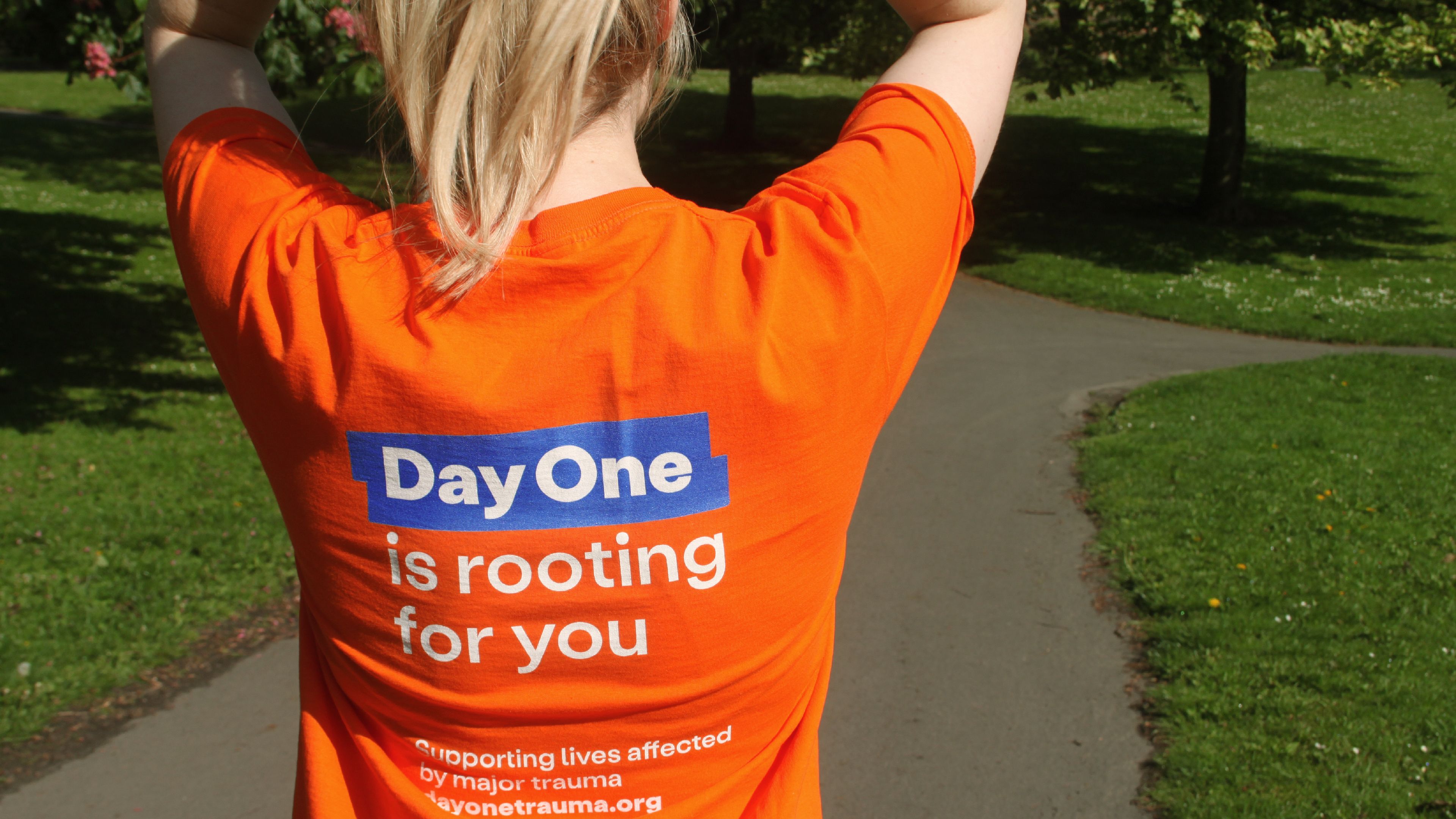 Image of a fundraiser wearing a Day One shirt is seen in a close up at the park. The writing on the back of her shirt reads: "Day One is rooting for you"