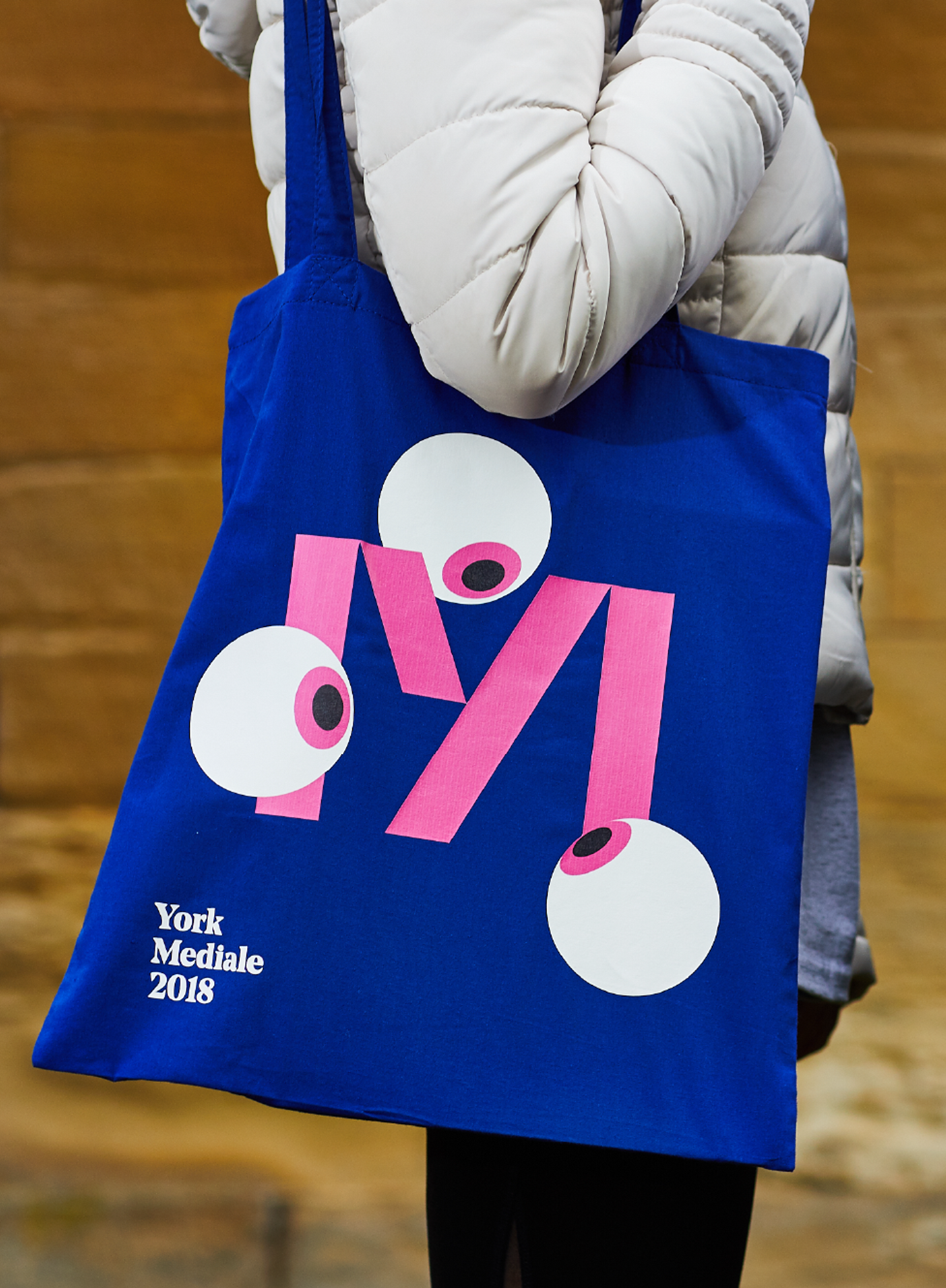 One person wearing a winter puffer coat holding an electric blue tote bag with the Mediale 'M' logo. Three eye balls surround the M, as if looking at it.