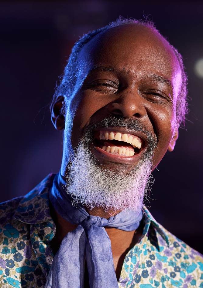 Elderly man wearing a funky shirt and knotted scarf is photographed mid laugh. The background setting is dark with purple lighting, that makes it look like it was taken from inside a club of some sorts. We are purely focussed on the man, as he's shot with a deep depth of focus