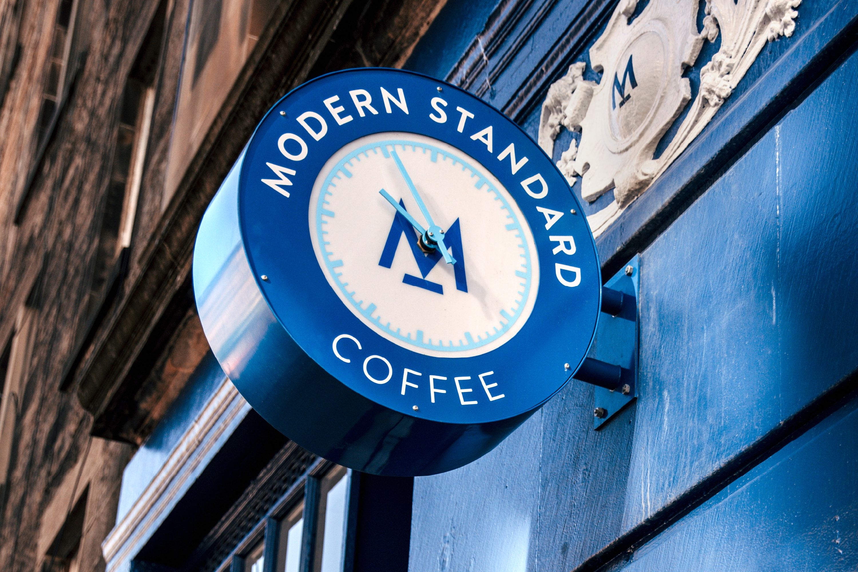 We see a round clock thats attached and protrudes from the outside of the Modern Standard cafe. The logo M is placed in the centre of the clock. The clock itself has light blue hands, as well as light blue indicators for the hour and minutes. Text surround the clock saying: "Modern Standard Coffee"