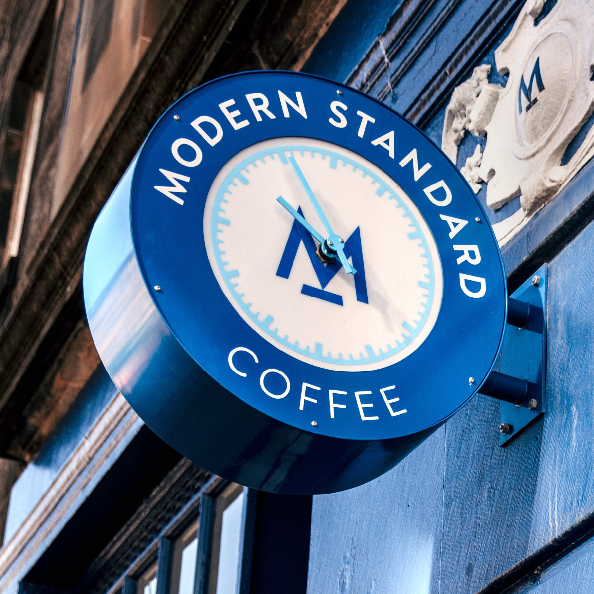 We see a round clock thats attached and protrudes from the outside of the Modern Standard cafe. The logo M is placed in the centre of the clock. The clock itself has light blue hands, as well as light blue indicators for the hour and minutes. Text surround the clock saying: "Modern Standard Coffee"