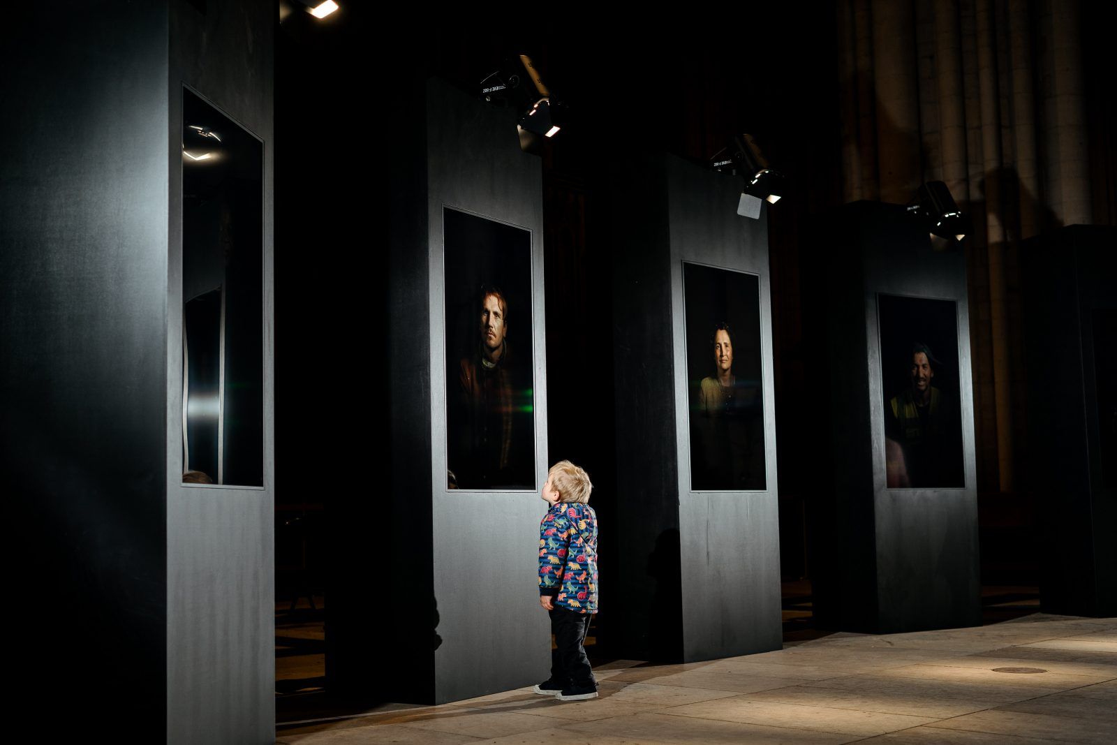 A child stands in a large gallery space looking up at one of the portraits on the wall. They are illuminated by the same light that shines on the portrait. Surrounding the child we can see four more portaits lit up, with the rest of the room shrowded in darkness