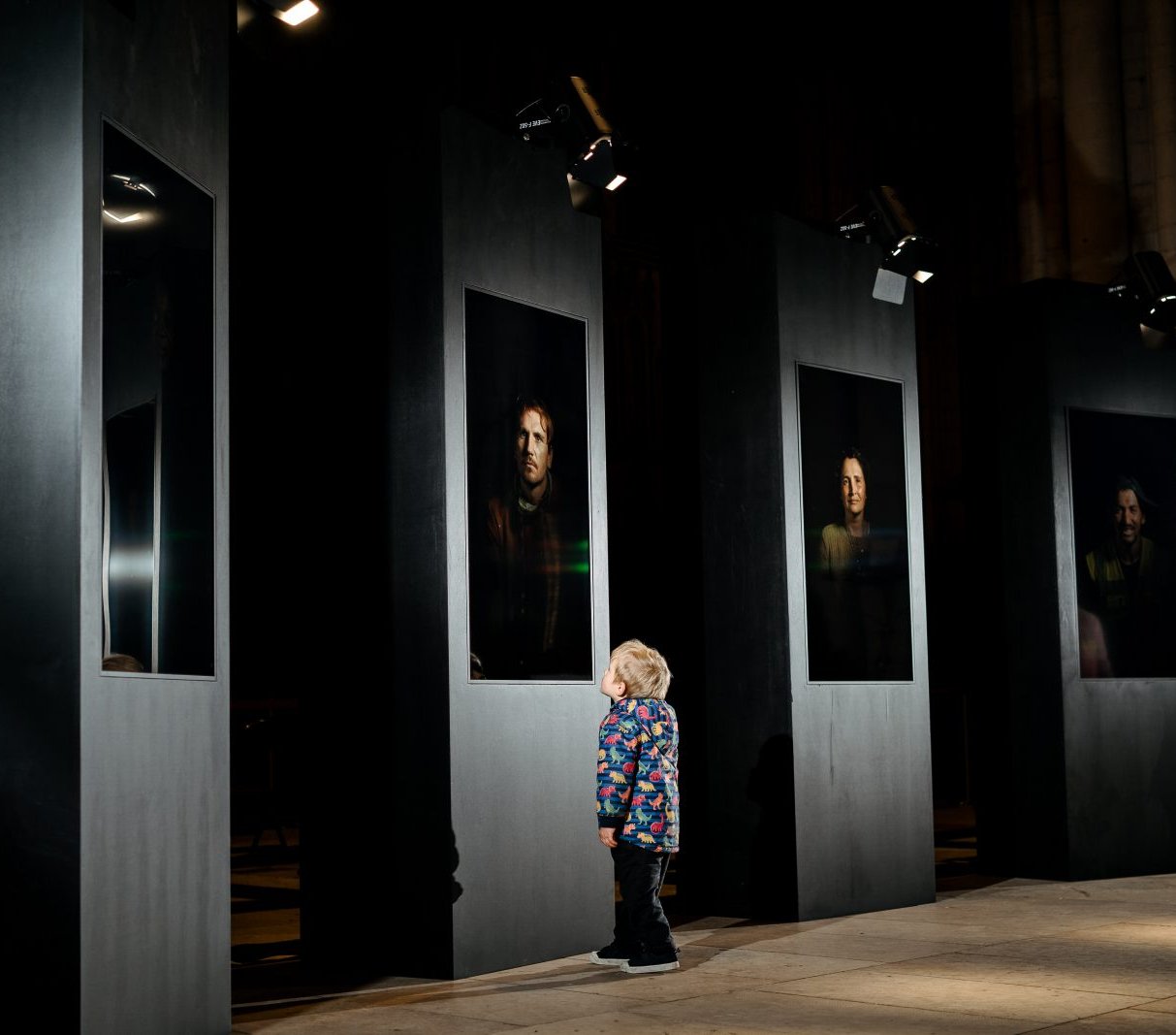 A child stands in a large gallery space looking up at one of the portraits on the wall. They are illuminated by the same light that shines on the portrait. Surrounding the child we can see four more portaits lit up, with the rest of the room shrowded in darkness