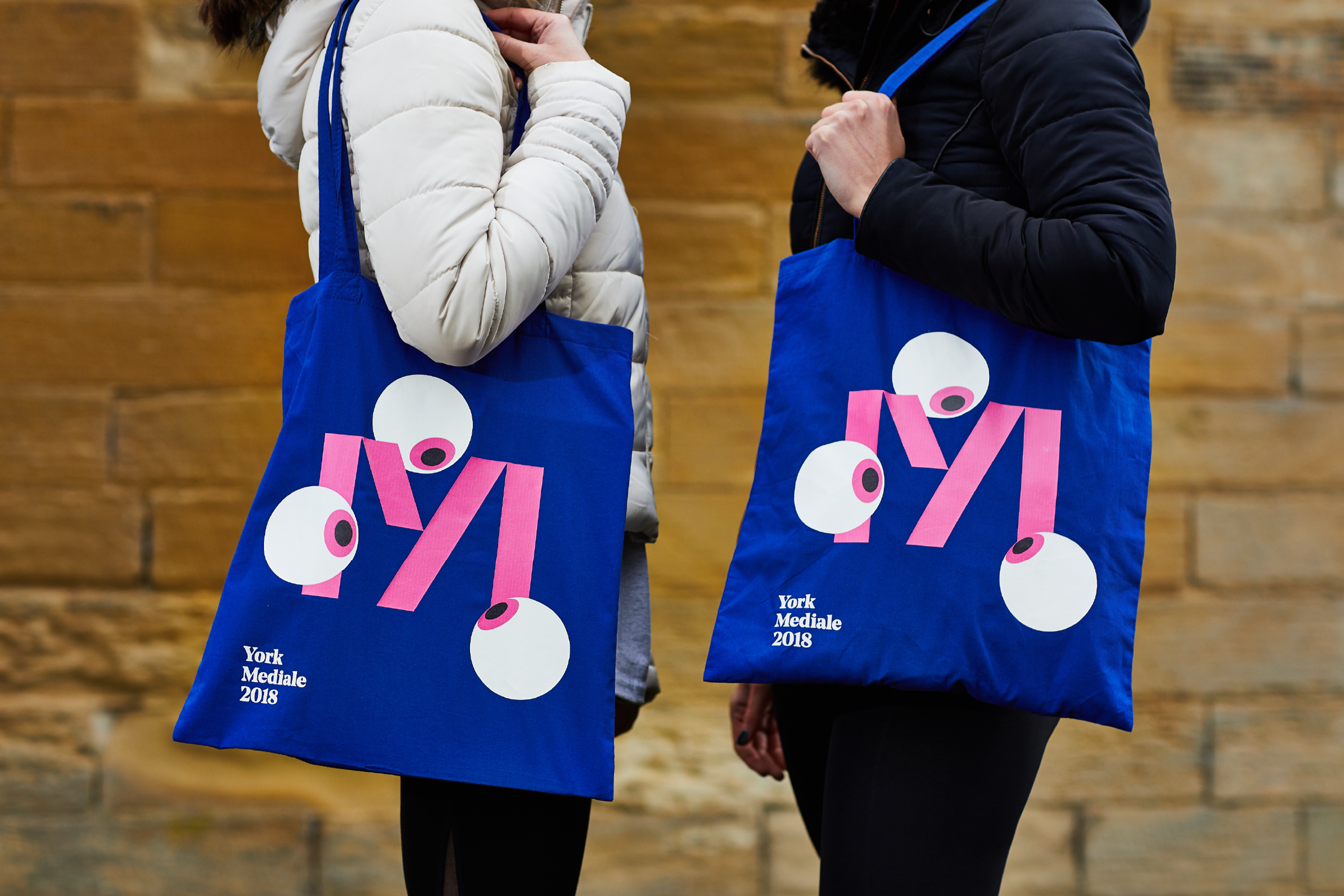 Two people wearing winter puffer coats hold electric blue tote bags with the Mediale 'M' logo. Three eye balls surround the M, as if looking at it.