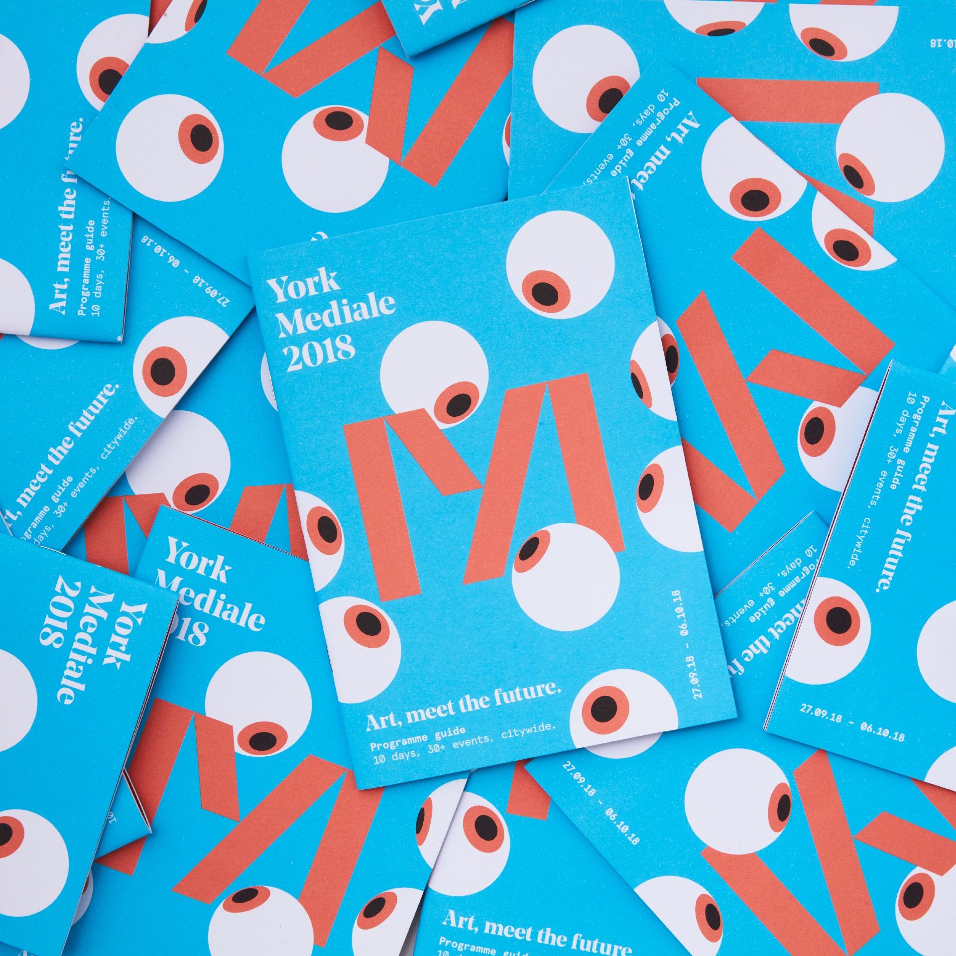 An overlapping pile of flyers for the 2018 Mediale is shown in a way that the whole frame is filled with them pointing in every possible angle and orientation. The flyers are a bright and light blue, with eyeballs looking towards the M in the centre. The tag line, logo and some date information is laid out surrounding the corners of the flyers