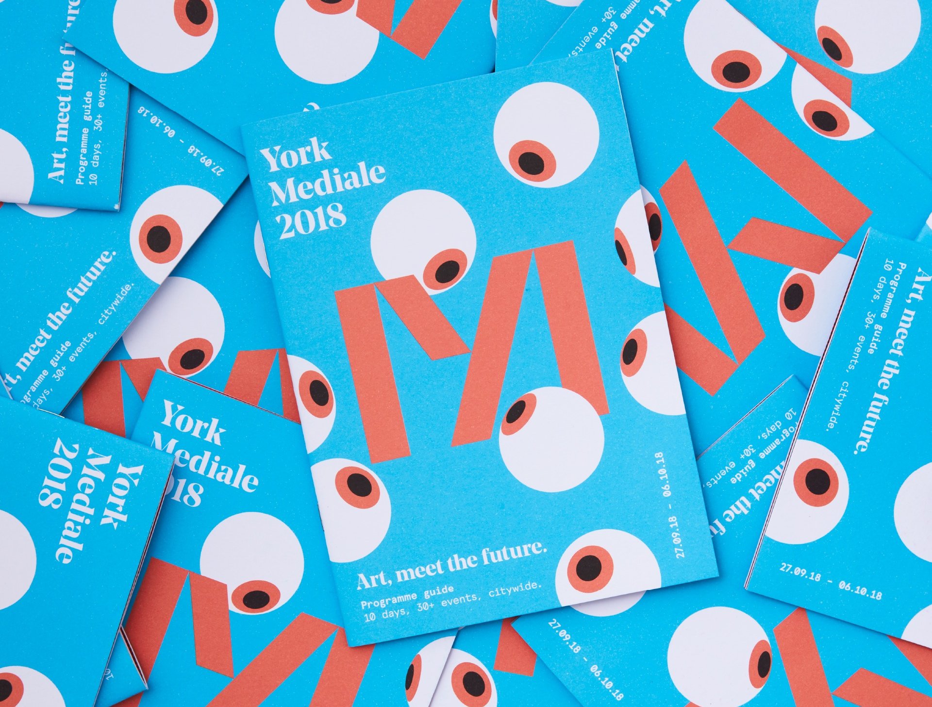 An overlapping pile of flyers for the 2018 Mediale is shown in a way that the whole frame is filled with them pointing in every possible angle and orientation. The flyers are a bright and light blue, with eyeballs looking towards the M in the centre. The tag line, logo and some date information is laid out surrounding the corners of the flyers