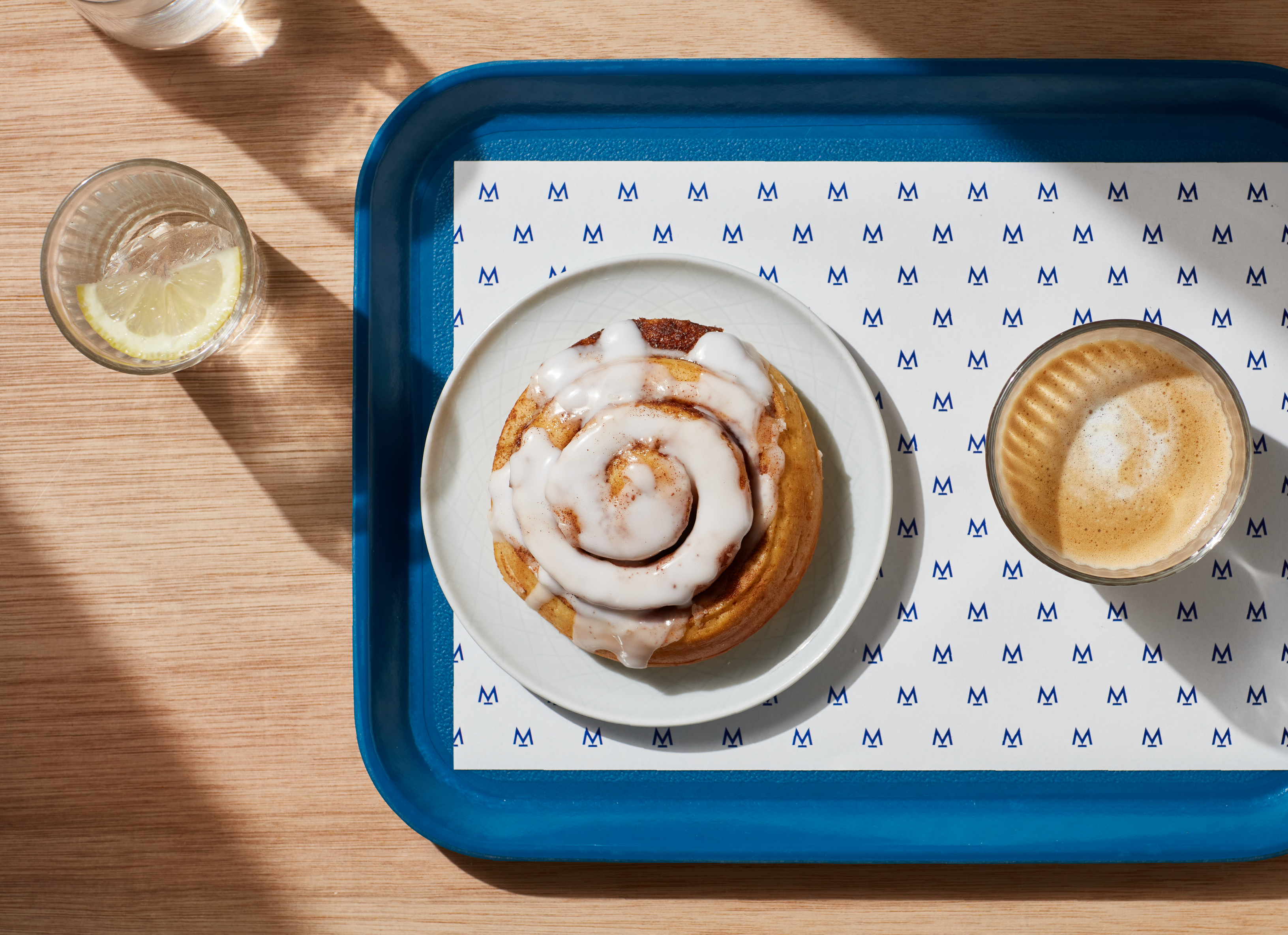 Woodern scandi style table, with a cup of water and lemon on the left. There is a blue tray on the right which fills 3/4th of the tray. On the tray is a sheet of Modern Standard Branding paper, cinnamon roll and accompanying coffee in a glass cup