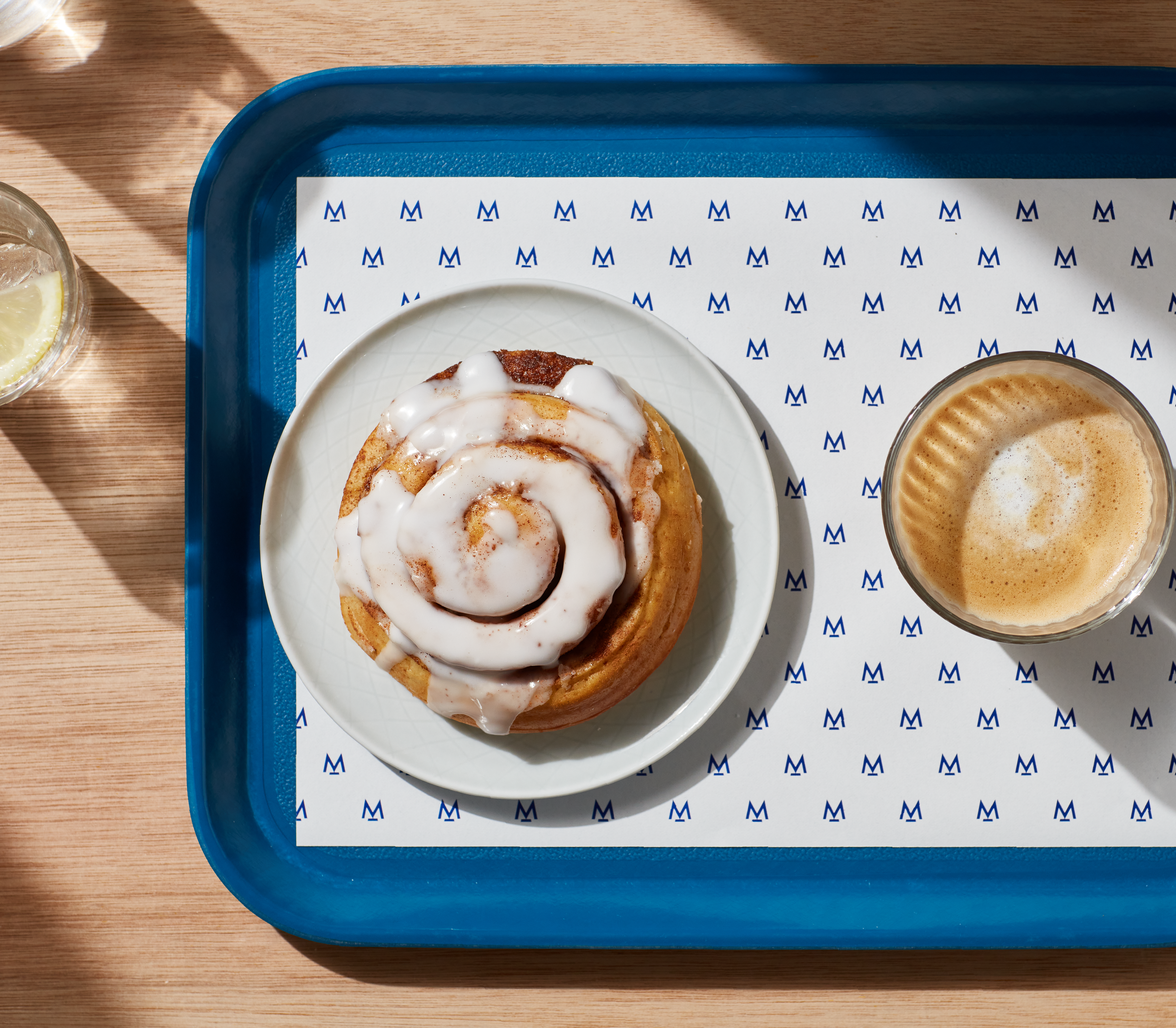 Woodern scandi style table, with a cup of water and lemon on the left. There is a blue tray on the right which fills 3/4th of the tray. On the tray is a sheet of Modern Standard Branding paper, cinnamon roll and accompanying coffee in a glass cup