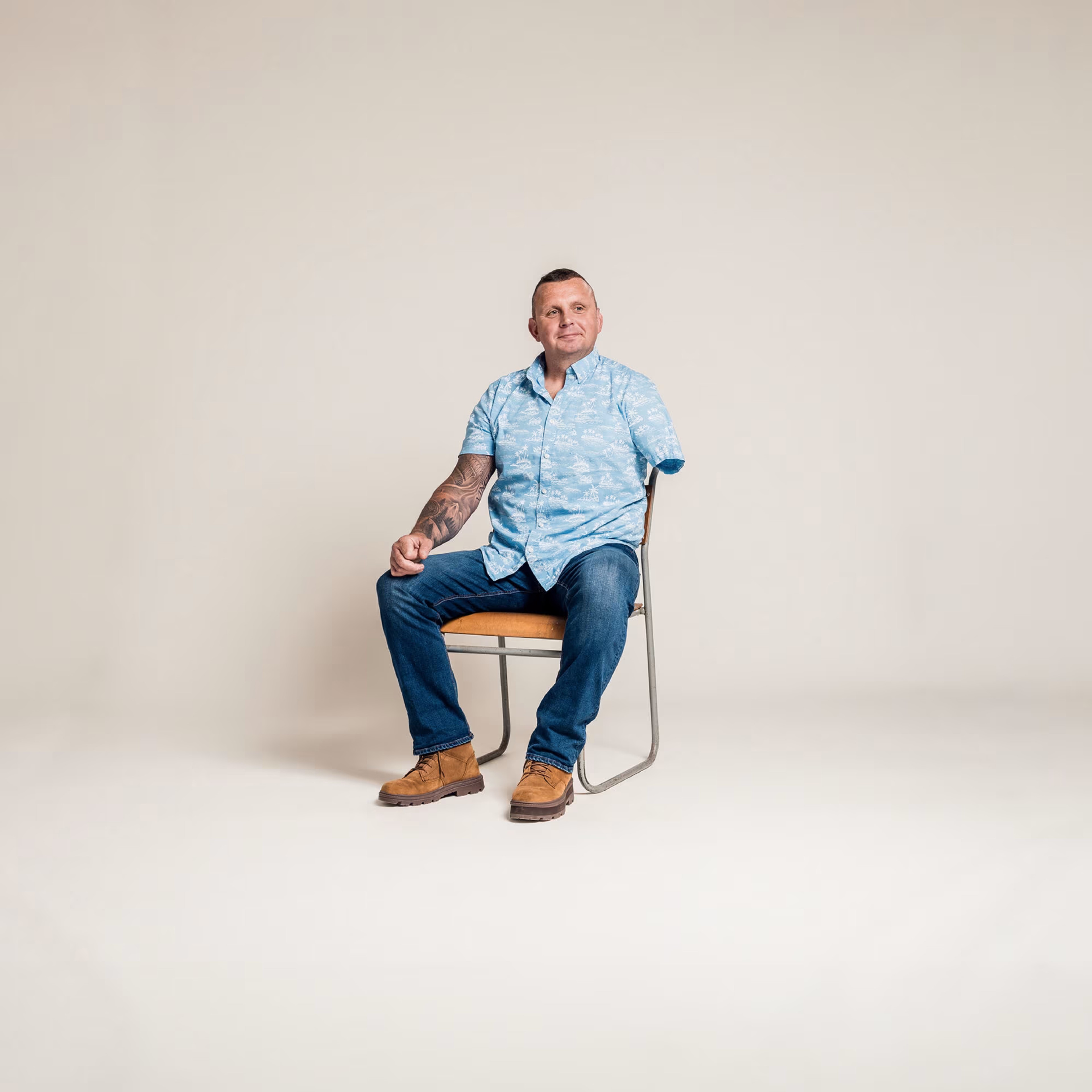 An amputee sits on a simple chair, with a cream coloured background. He wears a walm smile, and blue hawaiian shirt