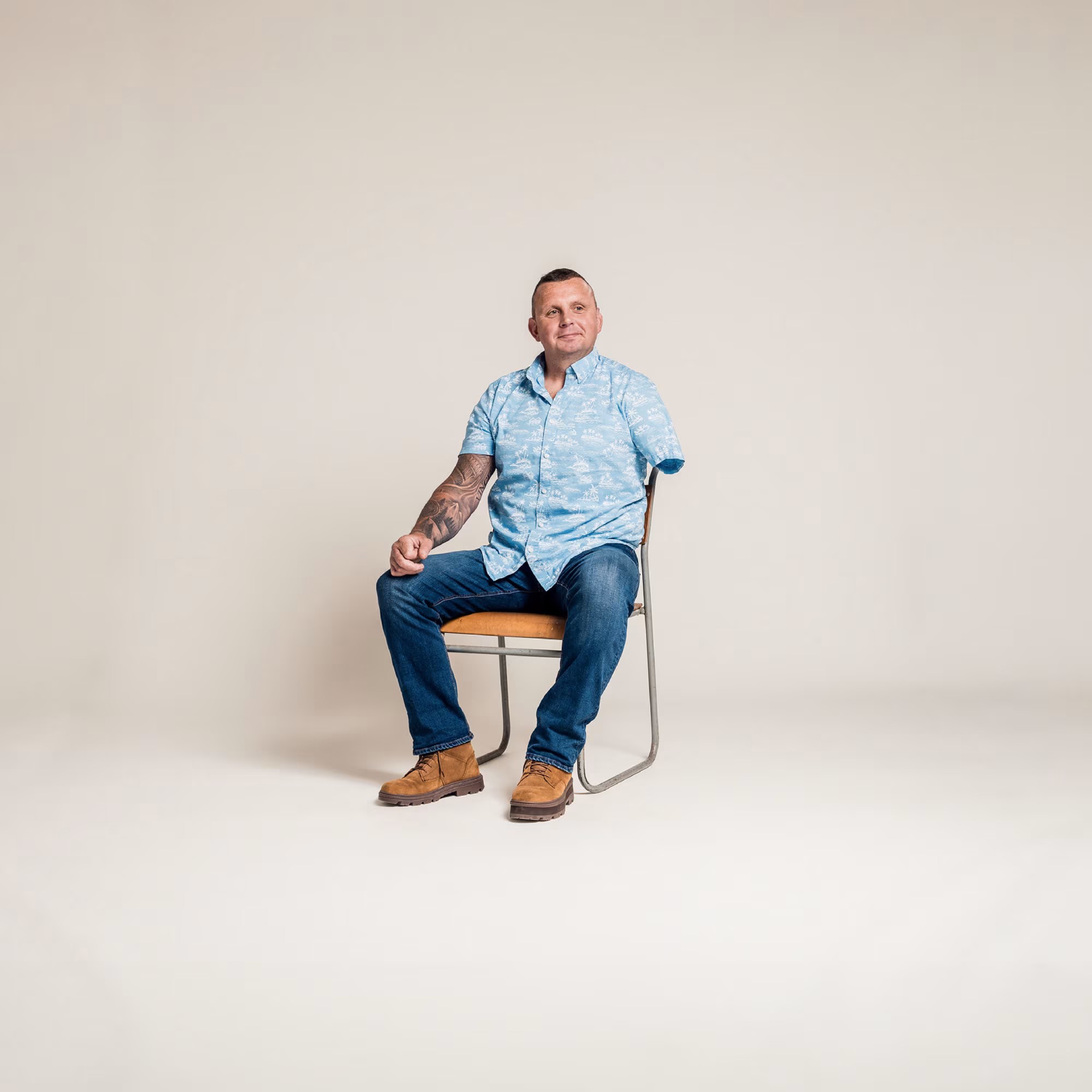 An amputee sits on a simple chair, with a cream coloured background. He wears a walm smile, and blue hawaiian shirt
