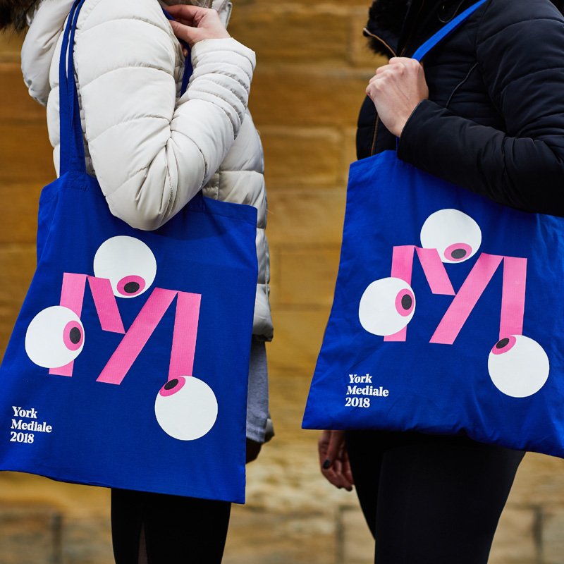 Two people wearing winter puffer coats hold electric blue tote bags with the Mediale 'M' logo. Three eye balls surround the M, as if looking at it.