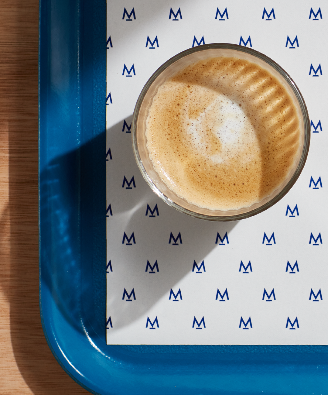 Woodern scandi style table is seen on the left, and a blue tray with a branding sheet of paper placed on the right. On this tray we have a freshly brewed coffee in a glass cup