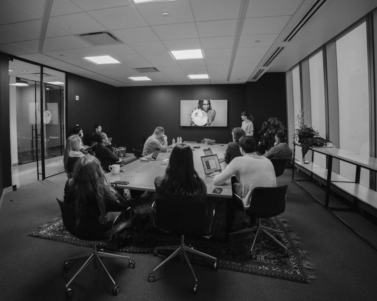 Black and white photo of a group of people in a conference room watching a presentation on a screen, illustrating a collaborative meeting environment.