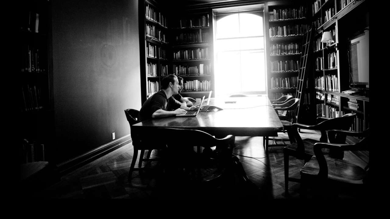 A man sits at a large table in a dimly lit library, working on a laptop. Bookshelves filled with books line the walls, and a large window provides natural light.
