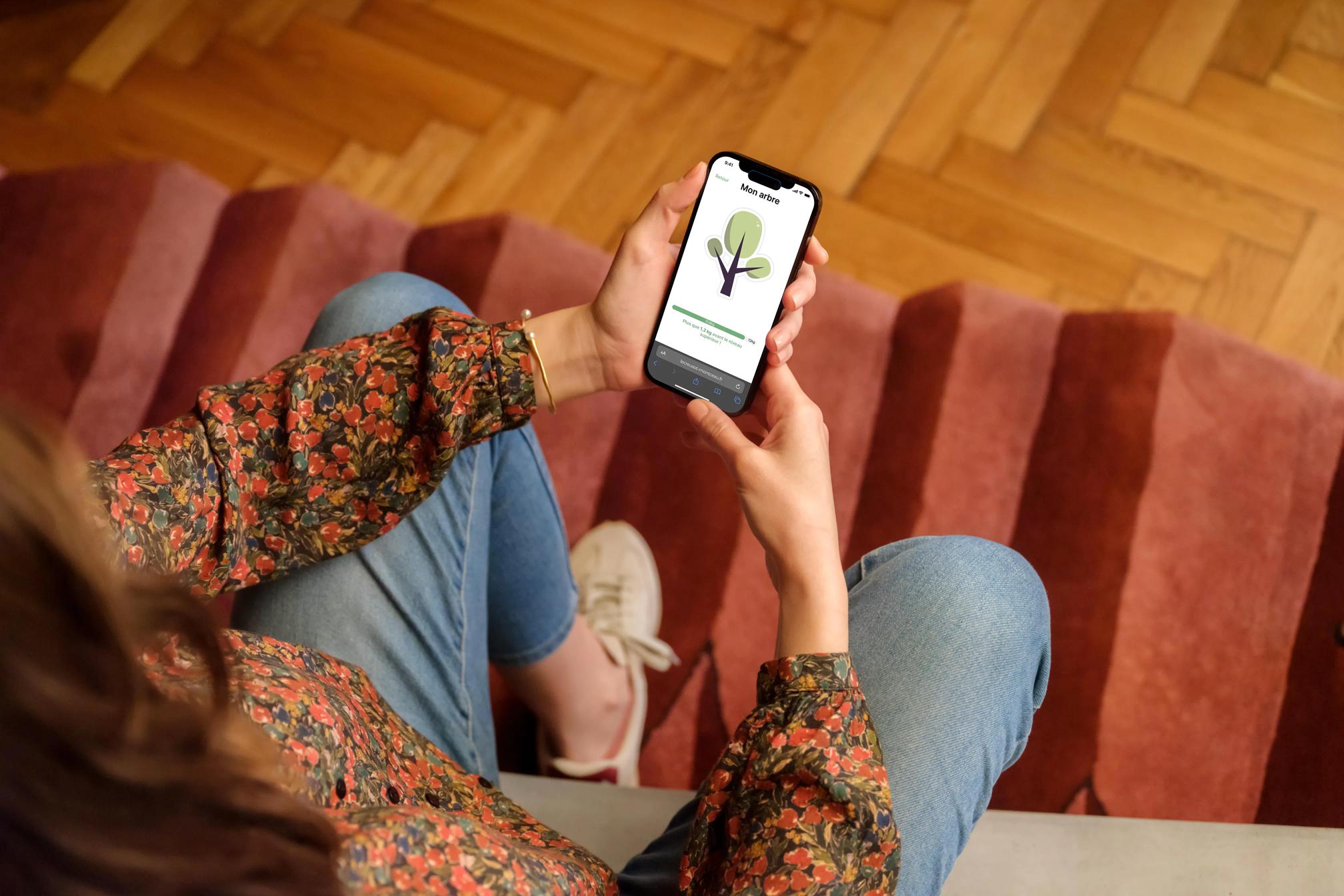 Une femme assise sur un escalier recouvert d’un tapis à rayures rouges, tenant un smartphone affichant une application mobile avec une illustration d’arbre et le titre ‘Mon arbre’ à l’écran. Elle porte une blouse à manches longues à motifs floraux et un jean.