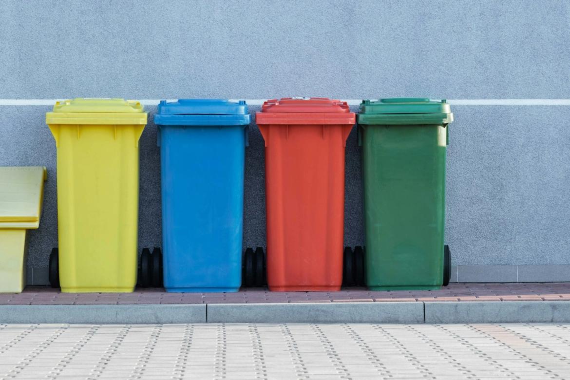 Quatre bacs de recyclage de différentes couleurs (jaune, bleu, rouge, vert) alignés contre un mur gris, représentant le tri sélectif des déchets.