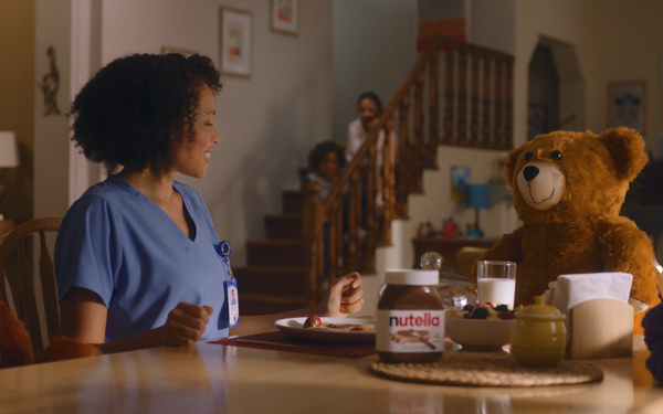 Woman enjoying breakfast with Nutella in a playful advertisement featuring a teddy bear at the table.