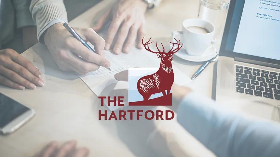 The Hartford Business Insurance logo featuring a red stag, overlaid on a photo of people reviewing documents at a table with a laptop and coffee.