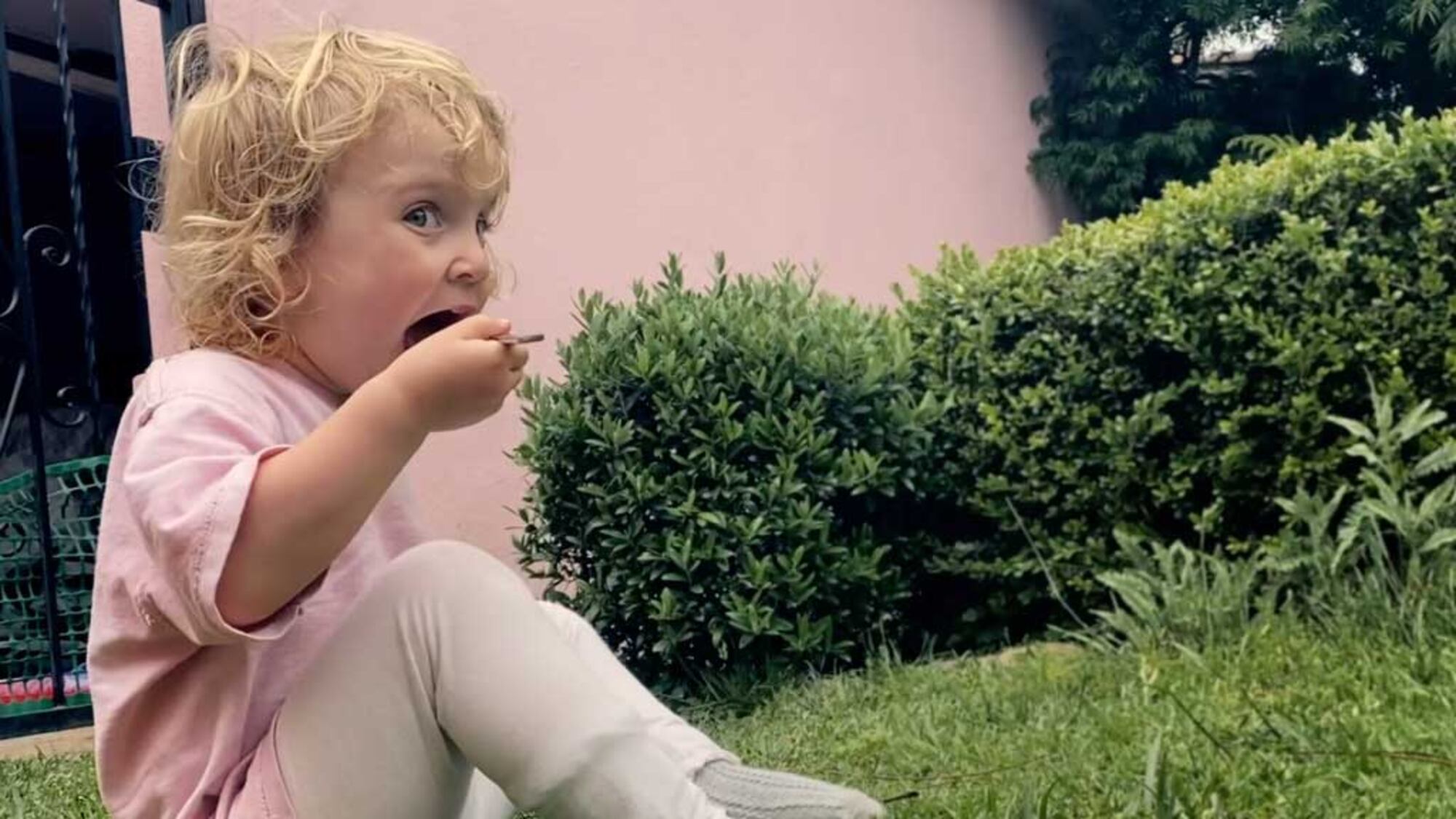 Young child eating with a spoon while sitting on the grass beside a garden hedge.