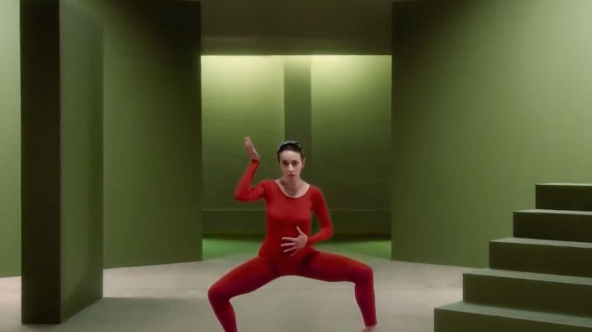 Woman in a red outfit dancing in a minimalist green room.