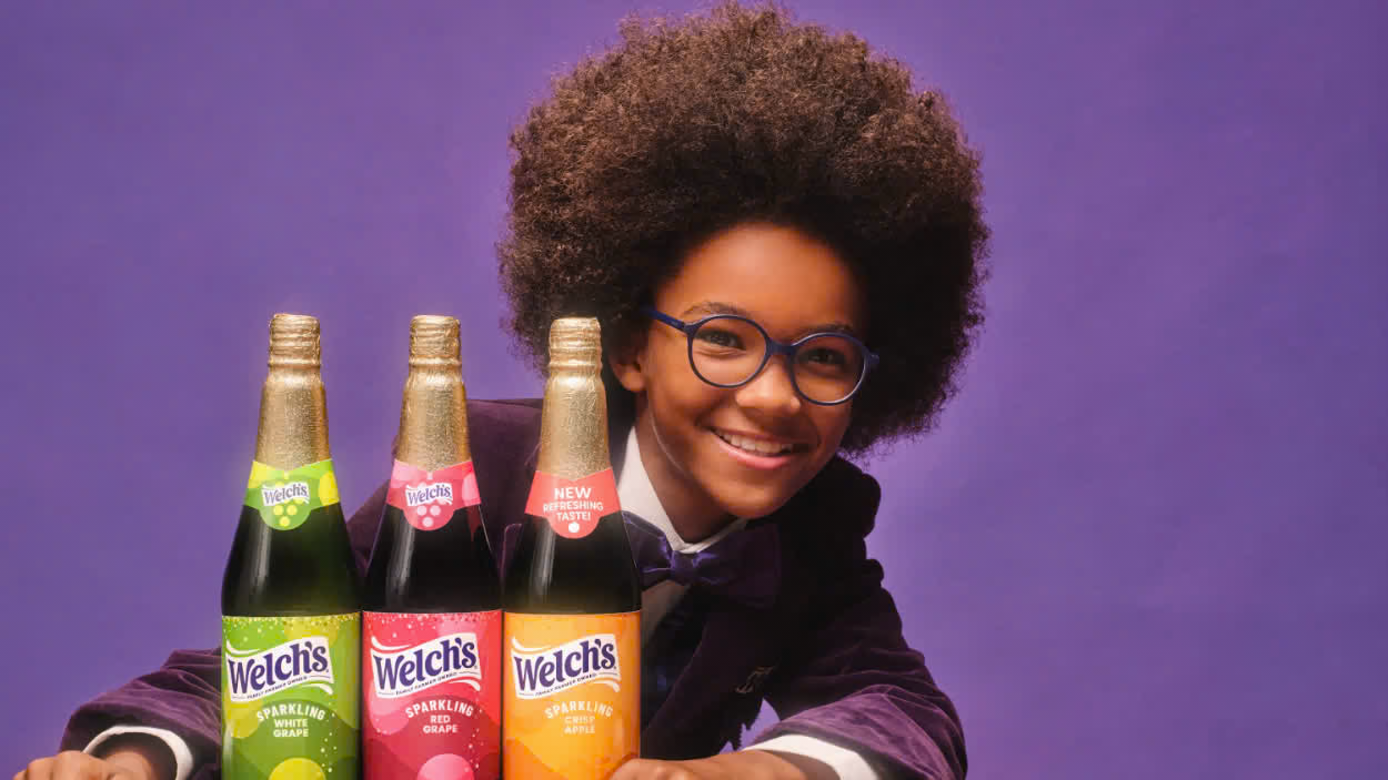 Young star wearing glasses and a purple suit poses beside three bottles of Welch’s sparkling juice in white grape, red grape, and crisp apple flavors.