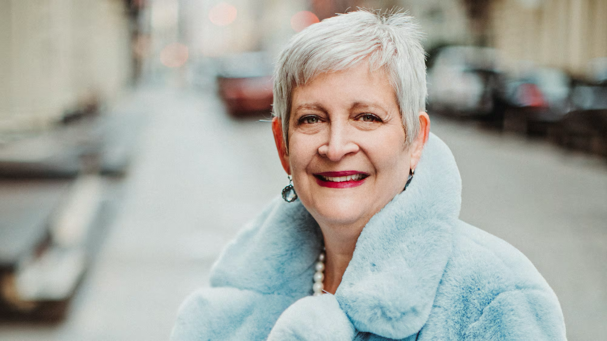 Portrait of Sandy Greenberg smiling in a light blue coat with a pearl necklace and earrings.