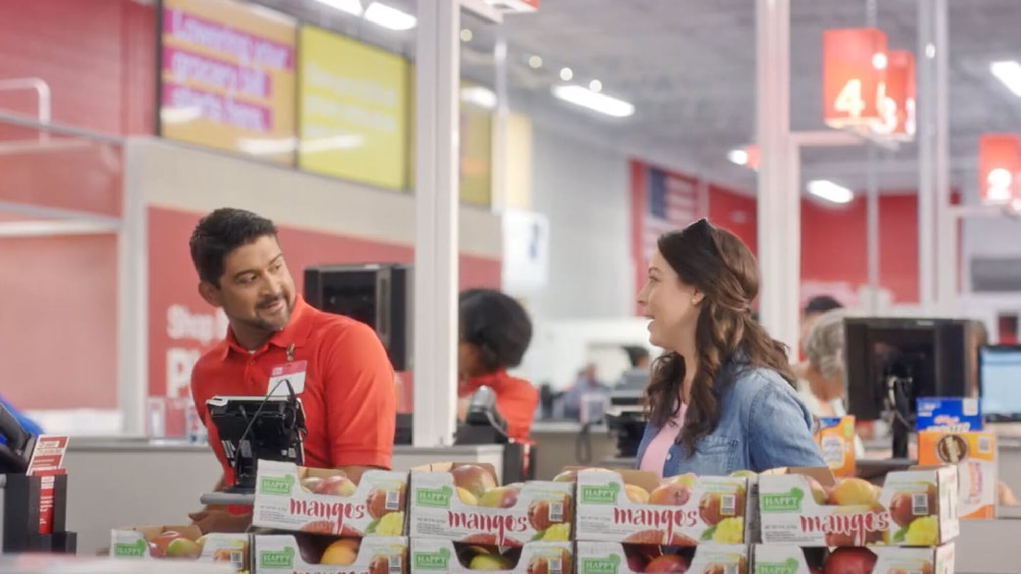 Grocery store employee in a red shirt chatting with a customer at checkout near displays of mangoes.