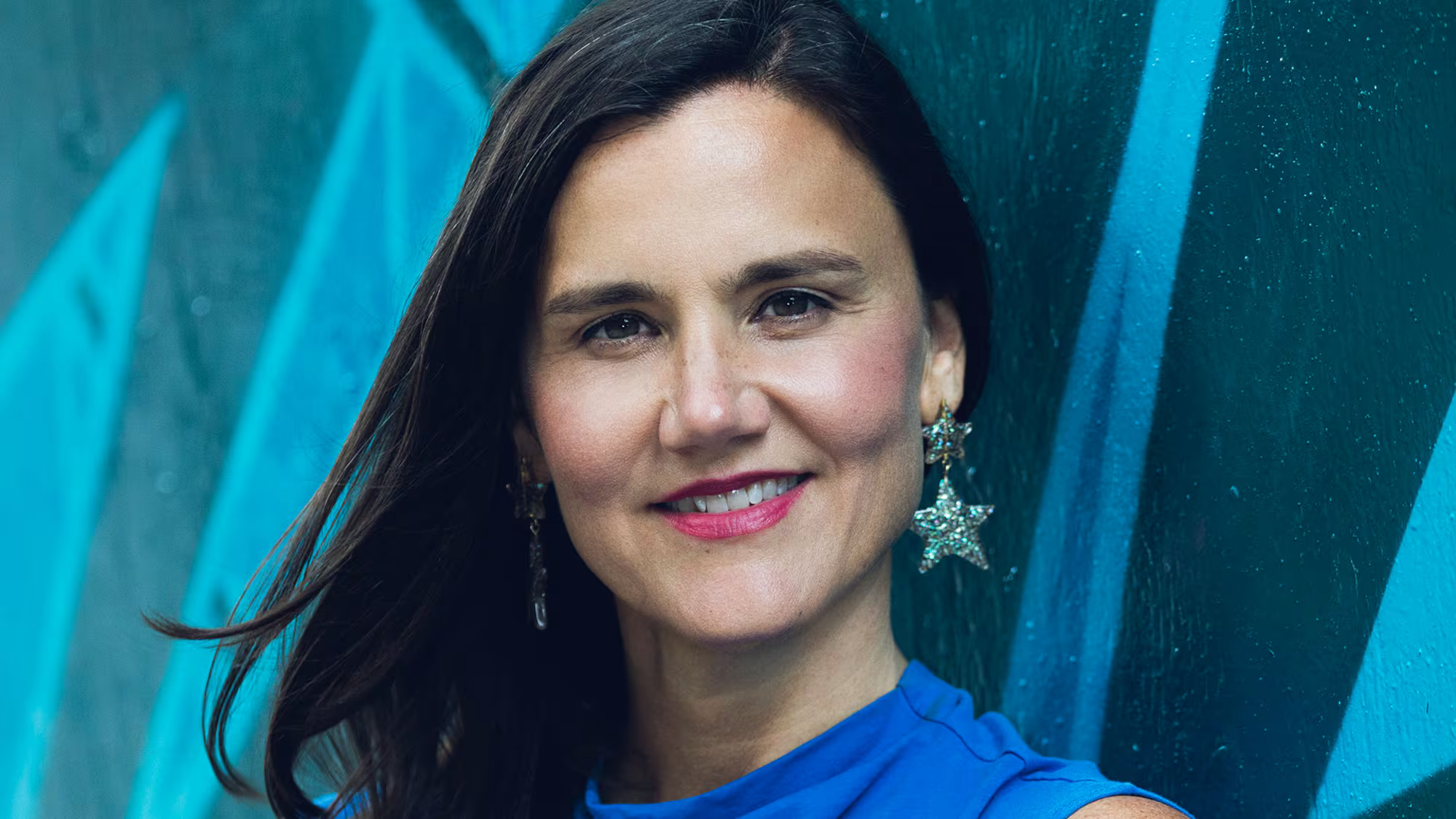Portrait of Terri & Sandy employee Amy Ferguson wearing a blue top and star-shaped earrings against a teal background.