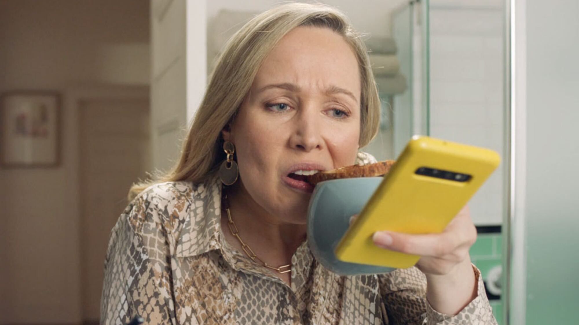 Busy woman holding a yellow smartphone while eating a slice of toast.