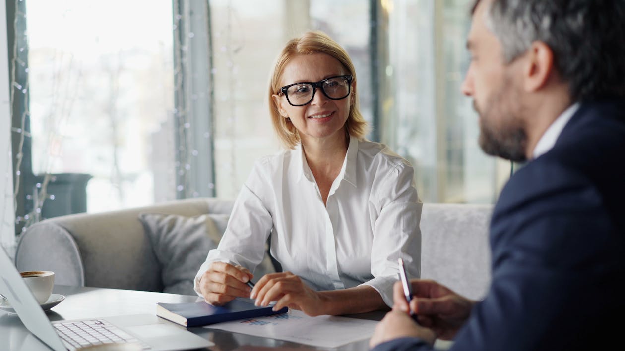 A smiling female professional in glasses listens attentively during a one-on-one consultation with documents and a laptop on the desk between them