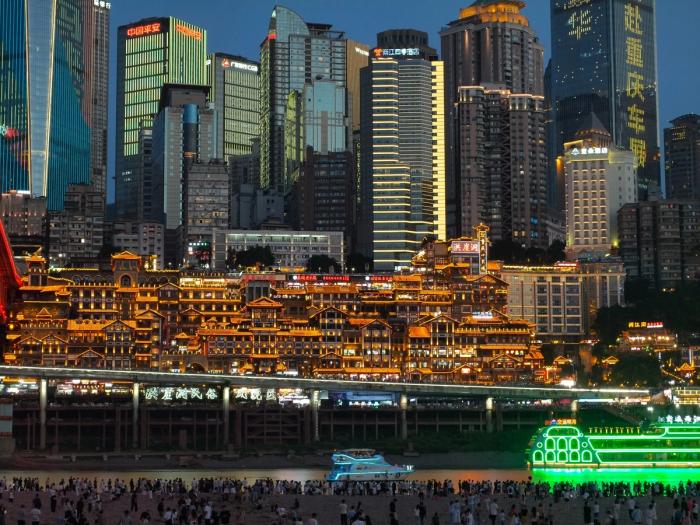 Blue-hour view across the river of the golden Hongyadong complex beneath Chongqing's high-rise skyline, with tour boats and crowds on the embankment