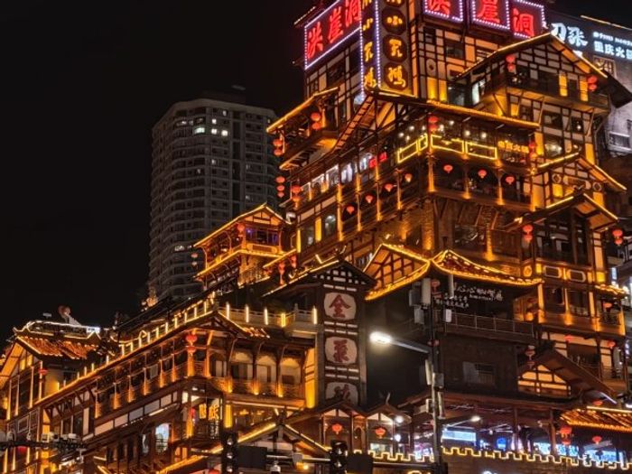 Hongyadong's stacked stilt buildings glowing gold at night, with neon 洪崖洞 signs on the top tower and a modern high-rise behind