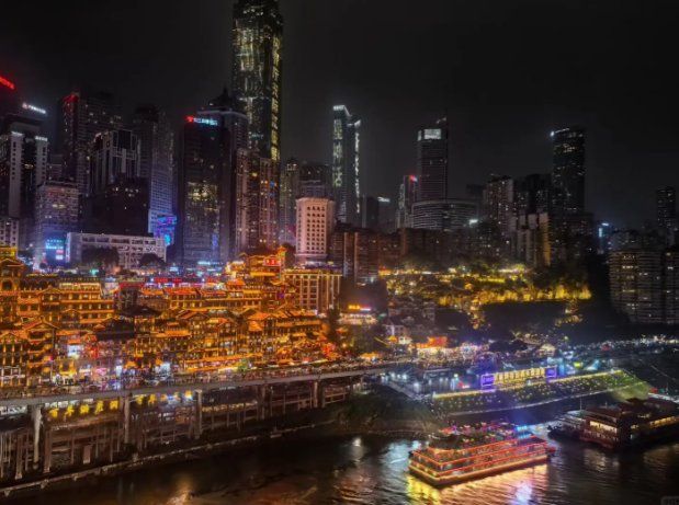 Wide night view of Hongyadong lit gold beside the Jialing River, with Chongqing's skyscraper skyline behind and illuminated tour boats on the water