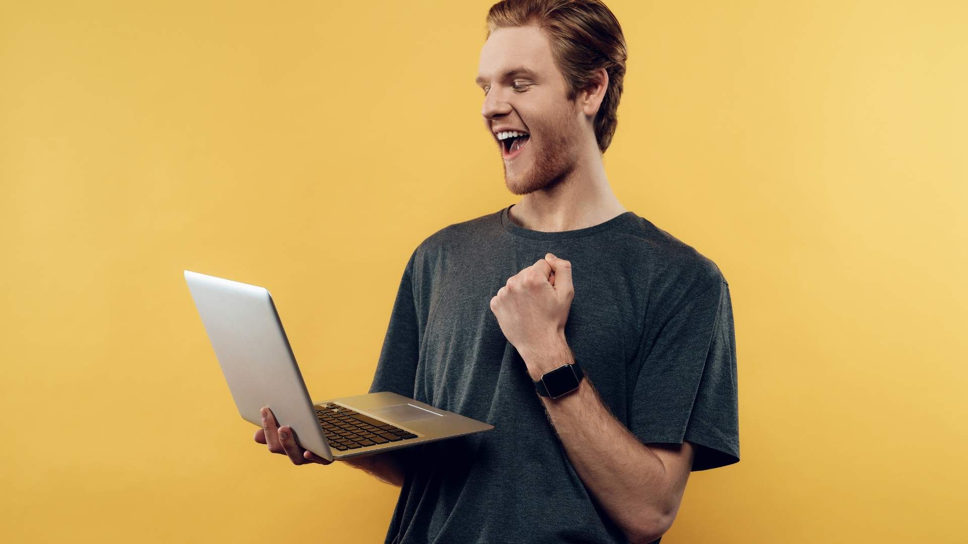 A person with short brown hair, wearing a dark shirt and holding a laptop in their hands while looking happy and proud.