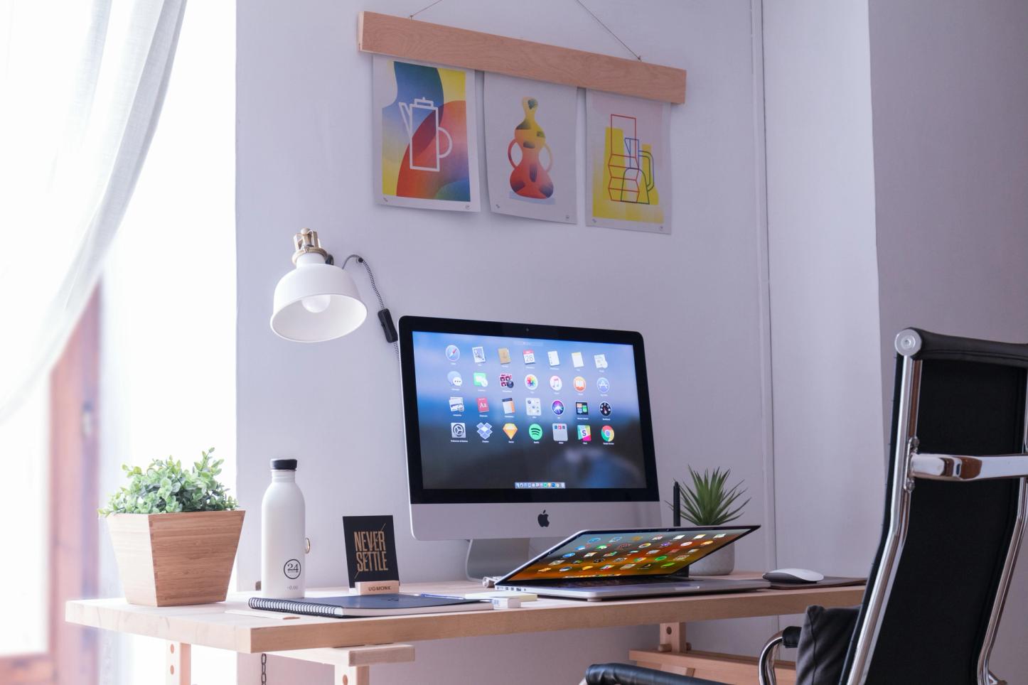 A home office set up with a wooden desk hosting an iMac with its screen showing an array of app icons. In front of the iMac, a MacBook lies flat and is partly closed, displaying a similar colorful screen. On both sides of the iMac is a small potted plant. On the right side is a white water bottle and a small black notebook. A white lamp is positioned to the left of the desk, behnd the iMac.  Three framed art prints hang above the the table on the wall. To the right, there's a black office chair with a chrome frame. The room has a cozy feel with soft natural light filtering through a curtain on the left, indicating a window just outside the frame.