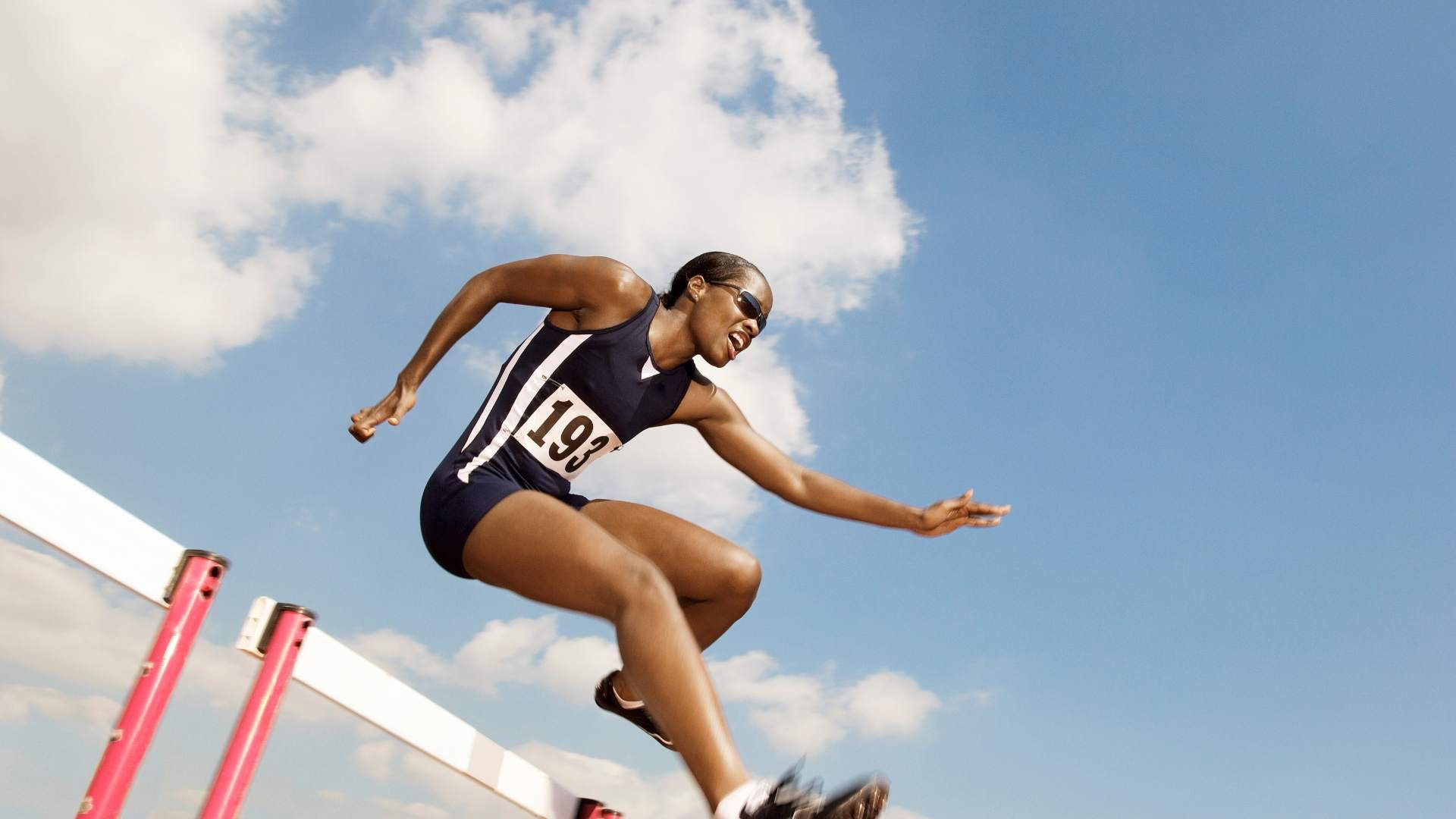 A Black person of color jumps over hurdles during athletics. The camera is at a low-angle shot.