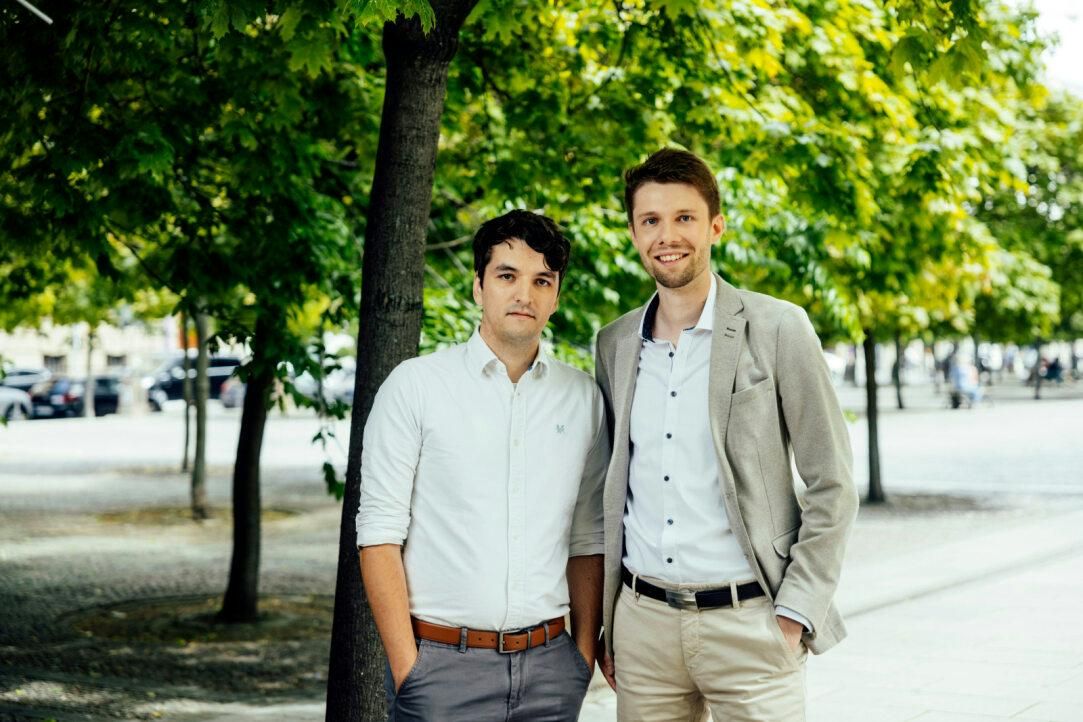 Benedict Padberg and Guido Zuidhof standing next to each other in a public space with a few trees in the background. They wear busniess attire, and smile into the camera.