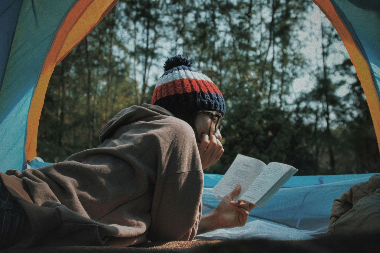 A person, wearing a red, white and blue beanie and a brown hoodie, laying on their stomach reading a book. They are in a blue tent, the front is open and the setting is in a forest.