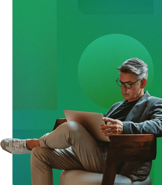 Man in glasses wearing business causal attire, crossing his legs, reviewing his DAM library on a laptop sitting in a chair with a geometric green background