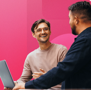Man wearing a sweater with glasses on holding his laptop explaining what digital asset management is to a man wearing a black button up shirt with a geometric magenta background