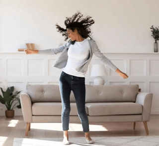Happy women dancing in the living room with her hands waving around with a grey couch in the background
