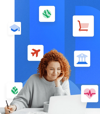 Women with curly red hair, working at a desk on a laptop holding a pen, leaning on her hands, with industry icons of higher education, healthcare, government, nonprofit and travel on a blue background